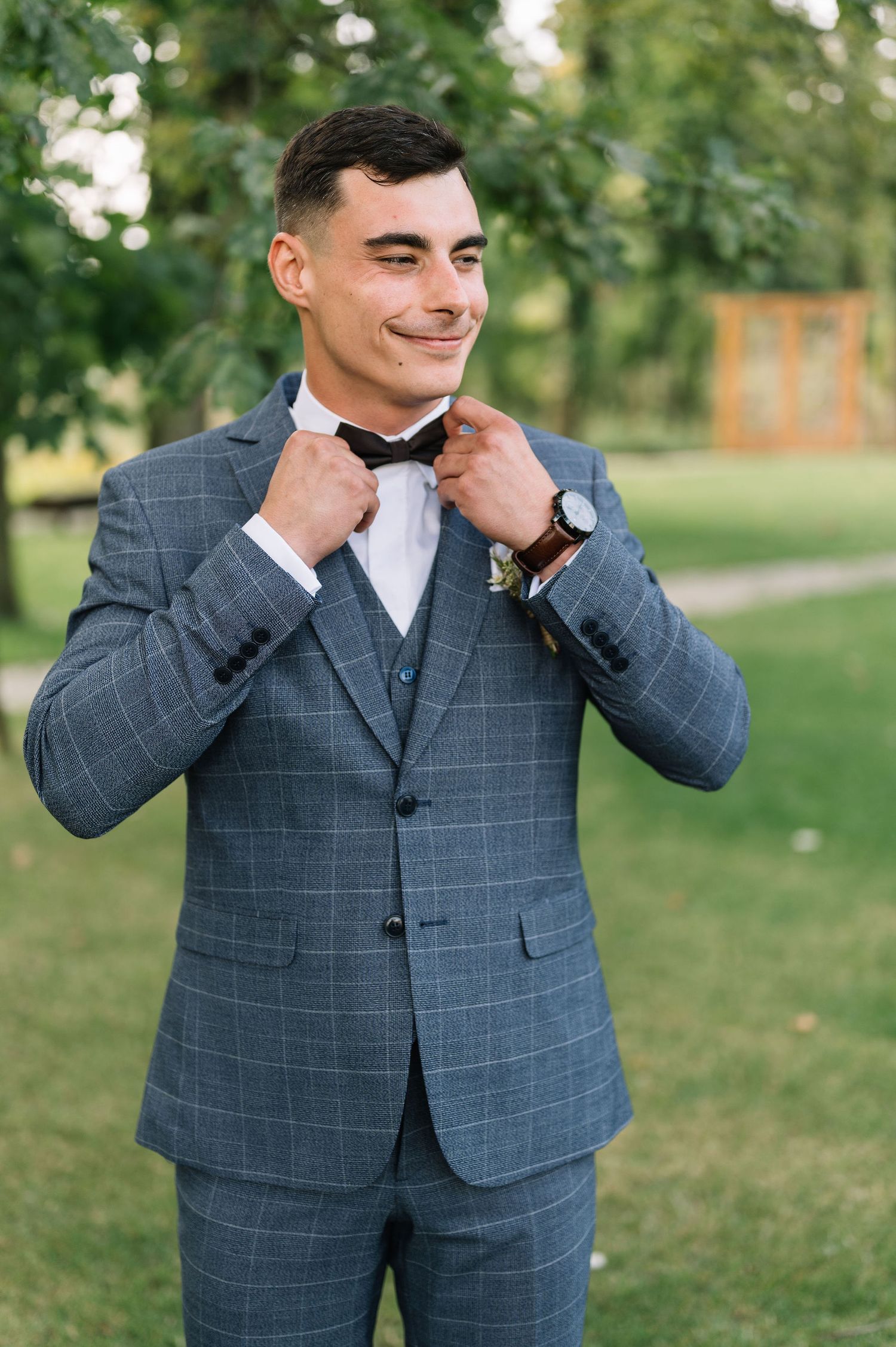 A groom adjusts his bowtie while wearing a grey plaid suit outdoors.