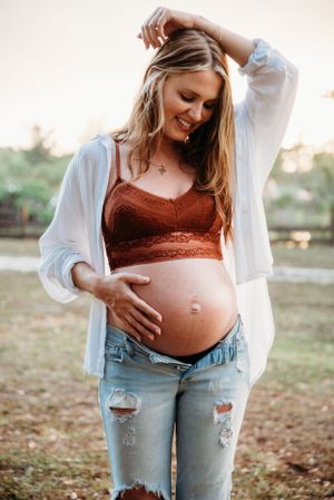 Expectant mother in distressed jeans and white cardigan poses outdoors during golden hour.