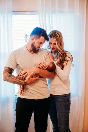 Parents in matching neutral colors holding their newborn baby by white curtains.