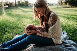 Mother in cozy sweater holding baby outdoors on blanket in green field.