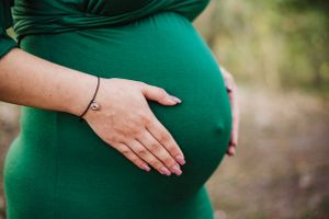 A pregnant figure in emerald green dress gently touching their belly outdoors.