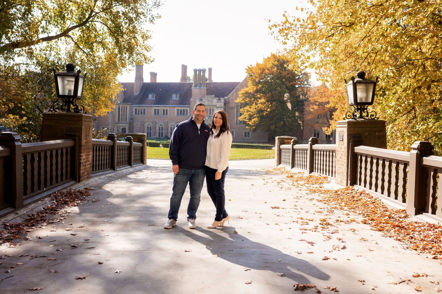 Couple walking along tree-lined path with historic mansion and autumn foliage in background.