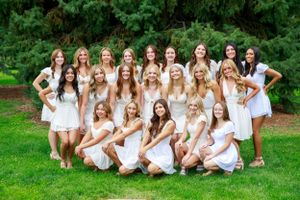Group photo of dance team members in white dresses posing on grass with trees in background.