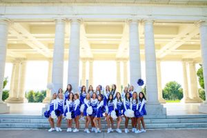 Cheerleading squad in blue uniforms poses on steps of classical building with columns.