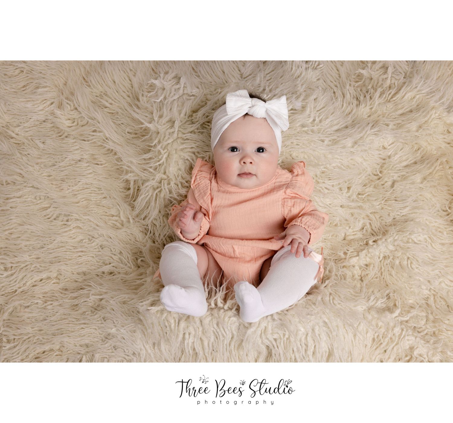 Baby wearing peach romper and white headband lying on fluffy beige rug during photography session.