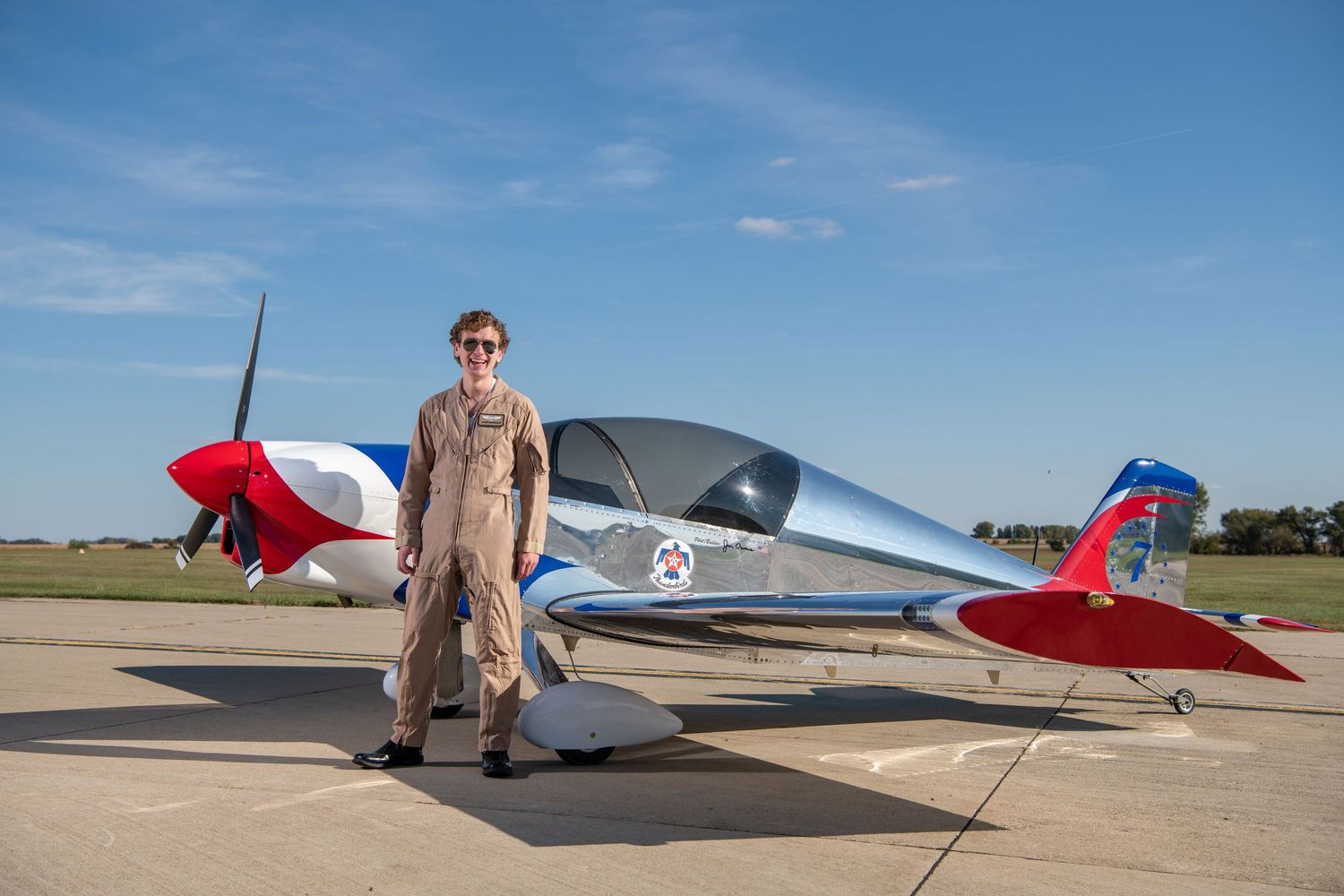Pilot stands proudly next to red and white aircraft on sunny airfield.