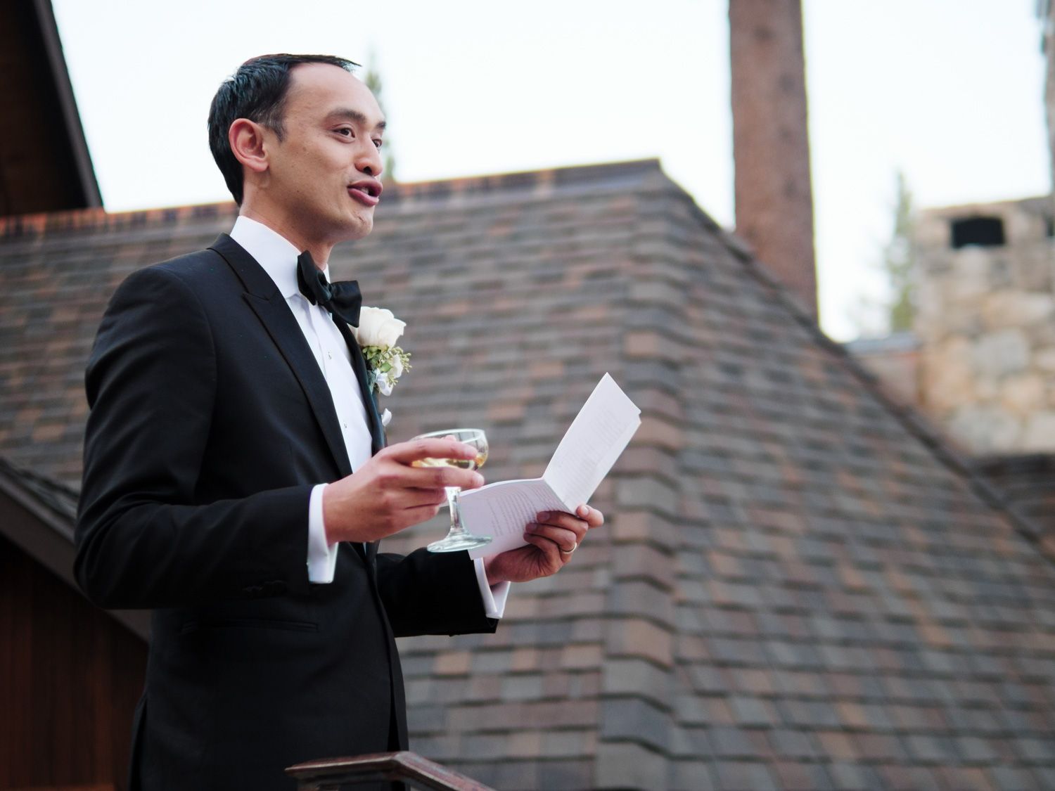 A person in a classic black tuxedo with bowtie reads from papers while wearing a white boutonniere against a wooden backdrop.