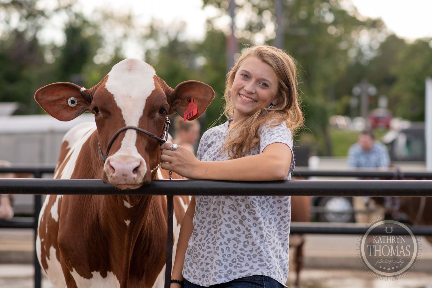 A brown and white cow posing with someone in a floral shirt by a fence at a farm event.