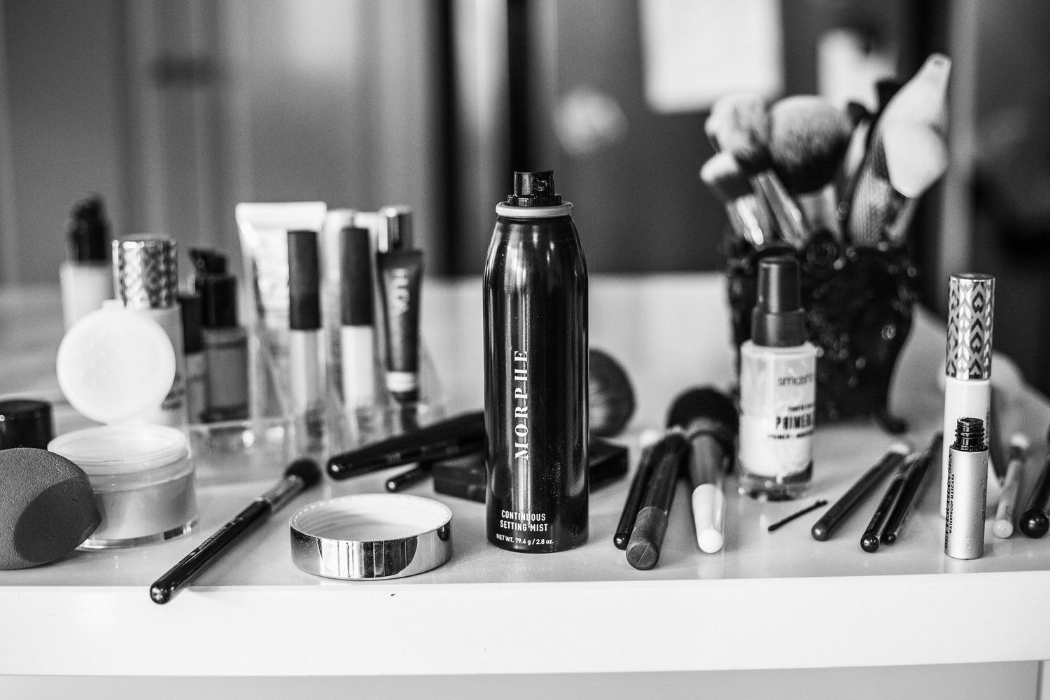 Black and white image of cosmetics and makeup brushes arranged on white vanity surface.