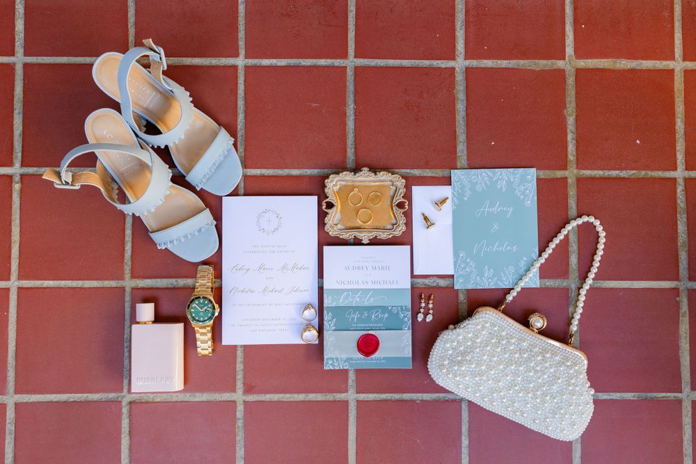 Wedding accessories including white shoes, invitations and a beaded purse arranged on terracotta tiles.
