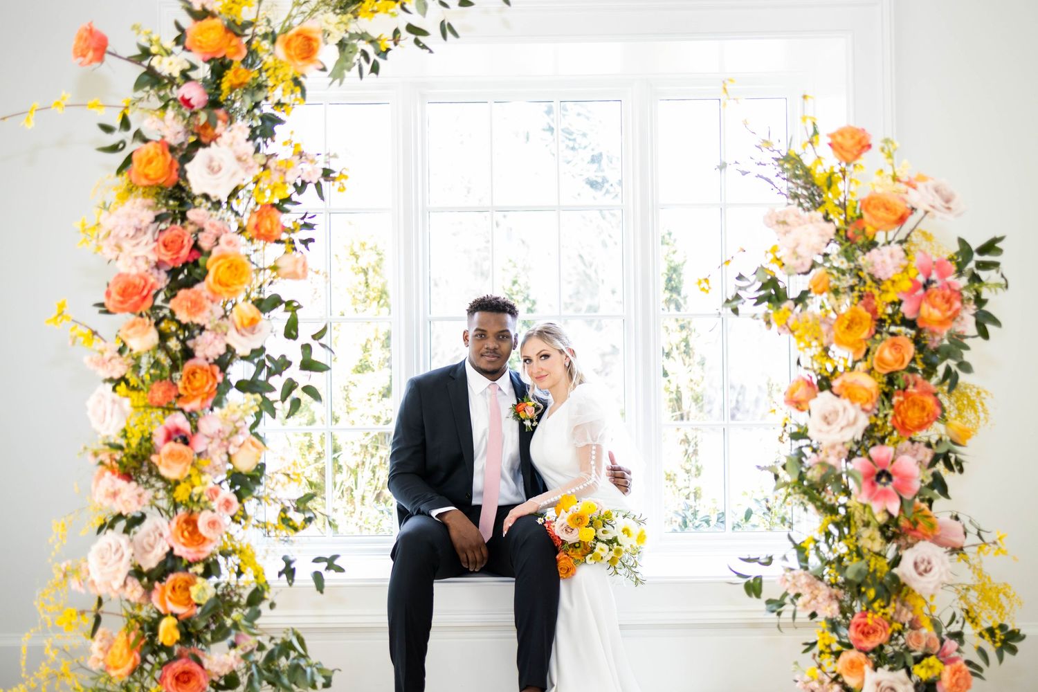 Bride a groom with floral arrangements surrounding a window with lots of natural light