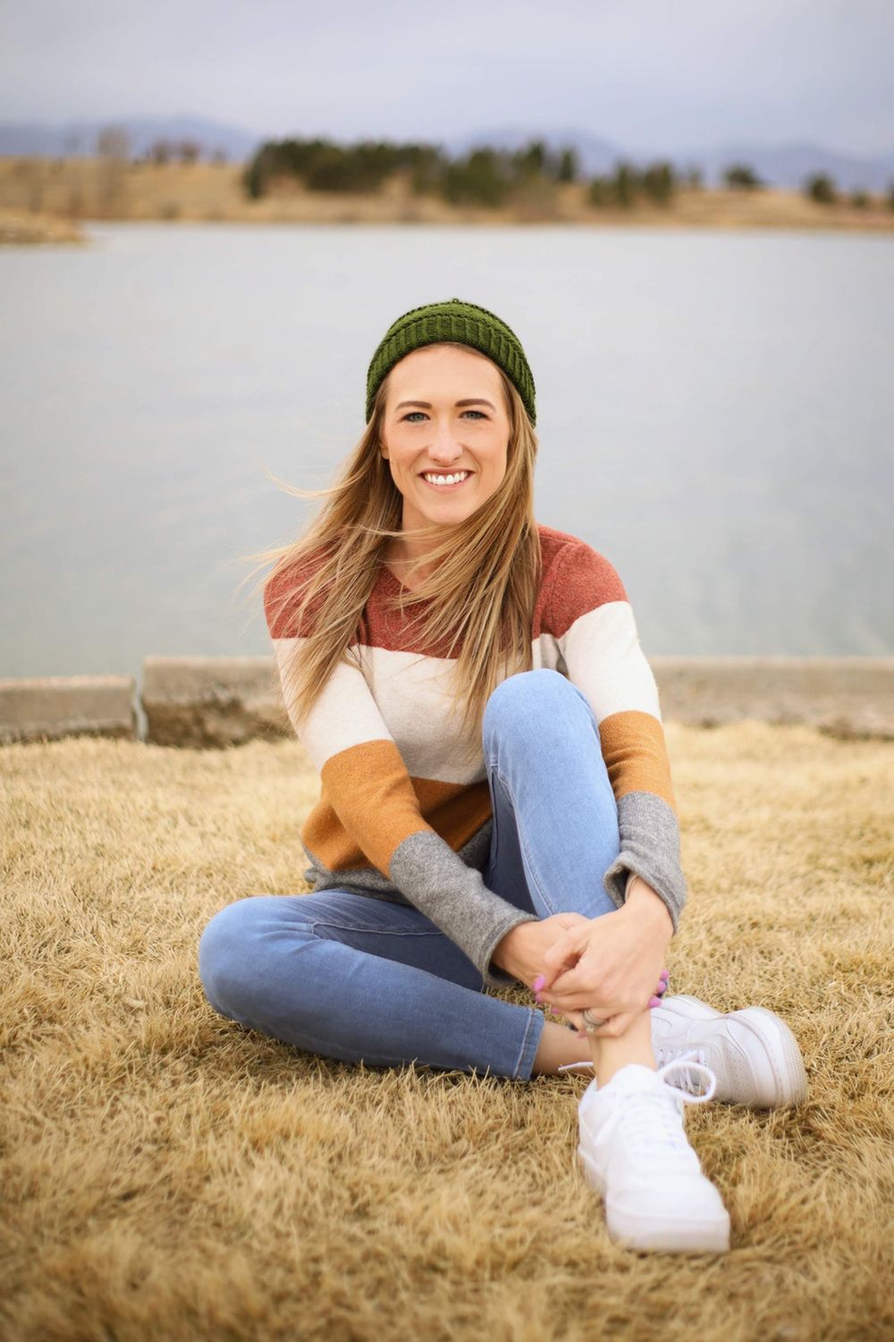 Person in colorblock sweater and green beanie sits on grass near lake wearing jeans and white sneakers.