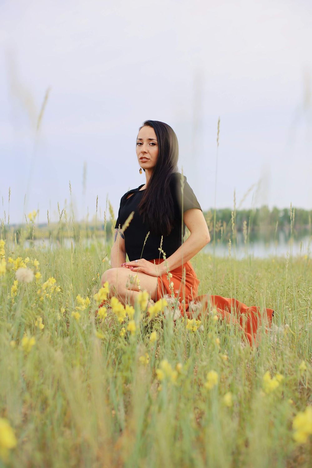 Person sitting in field of yellow wildflowers wearing black top and red skirt near water.