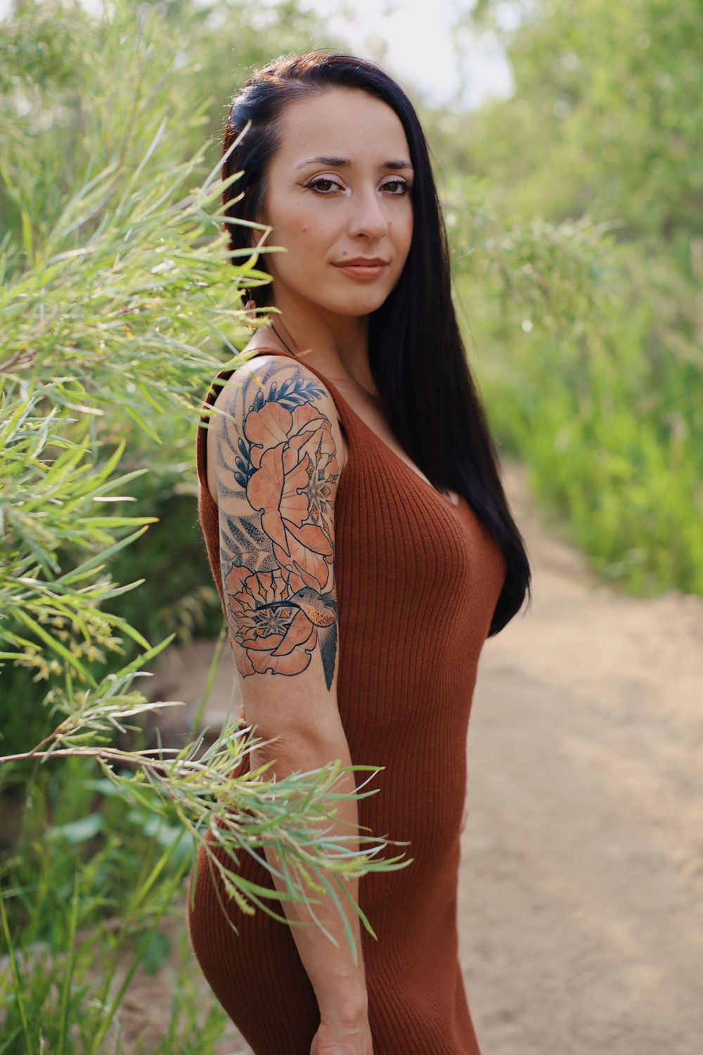 Person in brown dress shows floral arm tattoo while posing on nature trail.