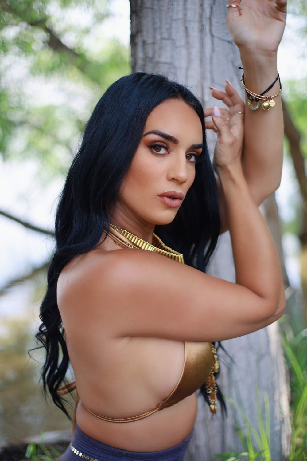Person wearing gold necklace and top poses against tree trunk with raised arms.