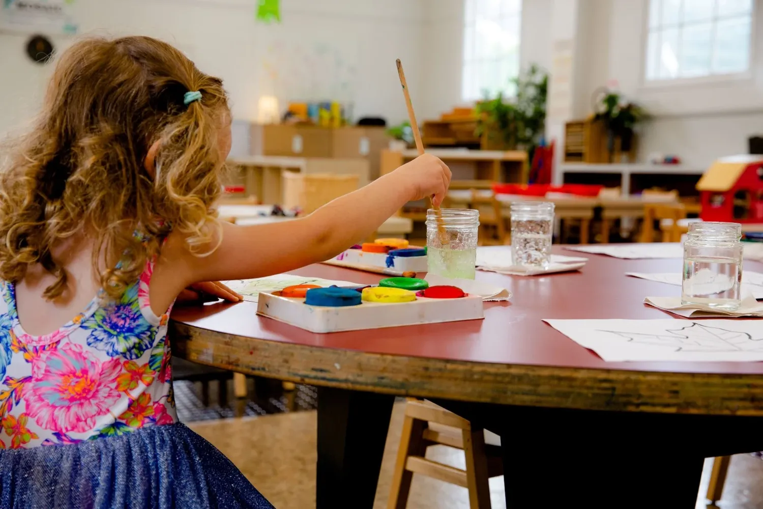 Student painting with watercolors at round table in bright classroom setting.