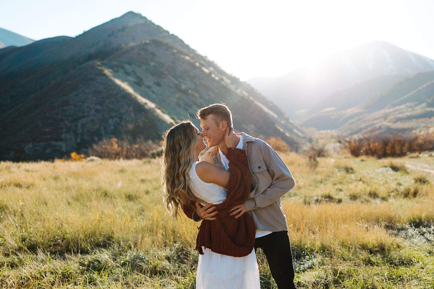 A couple embraces in golden sunlight with mountain peaks in the background.