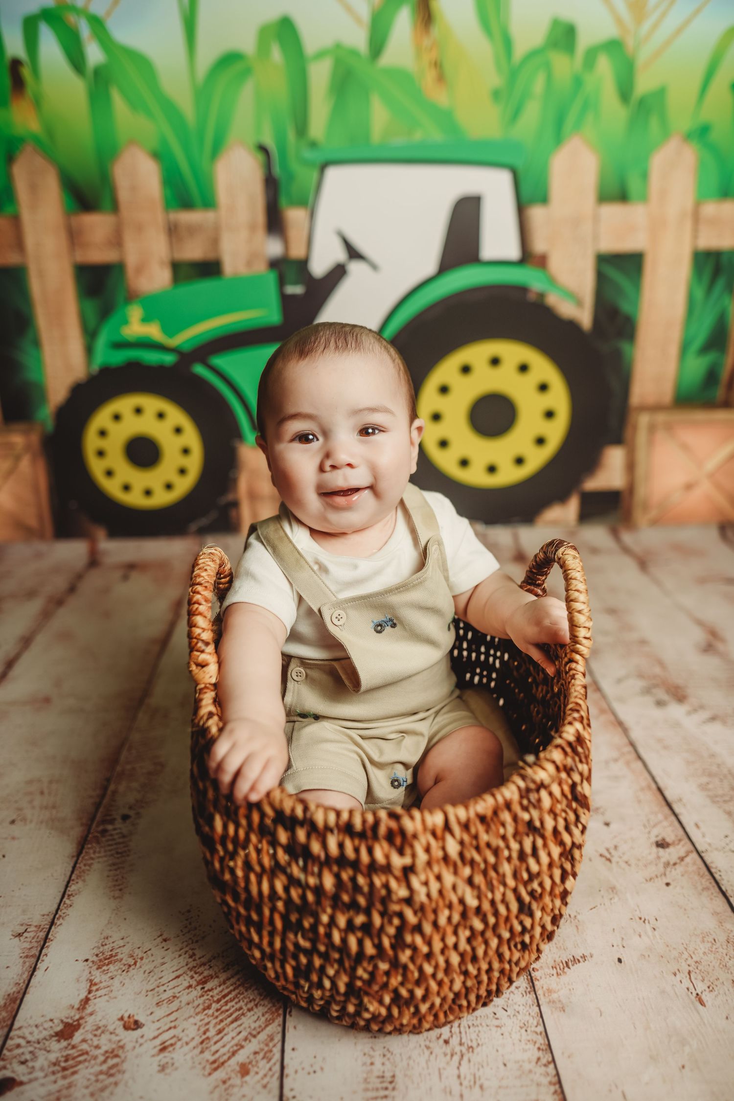 Baby in tan overalls sits in woven basket with green tractor backdrop and wooden fence.