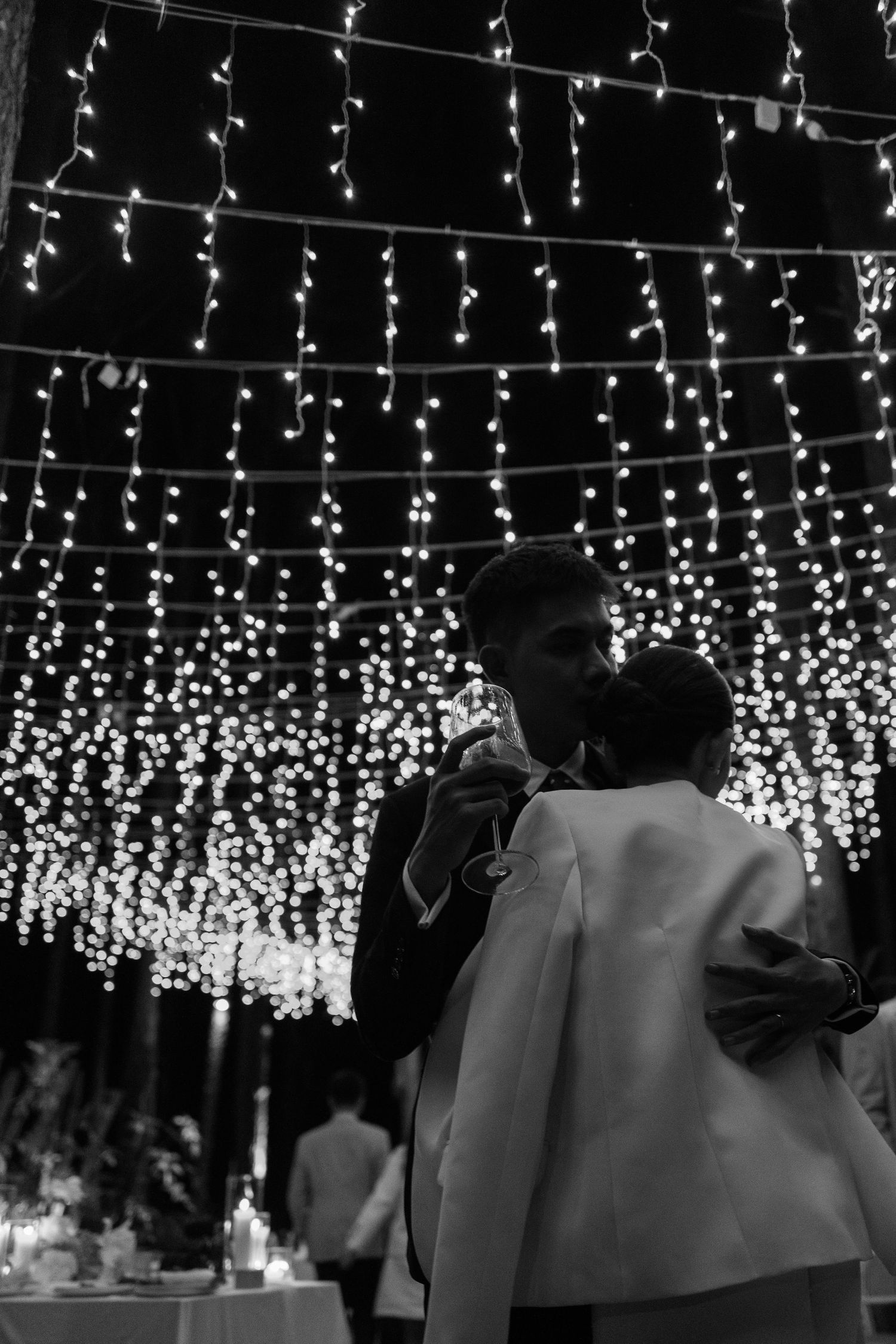 Black and white photo of bride and groom sharing a kiss under cascading fairy lights at their wedding reception.