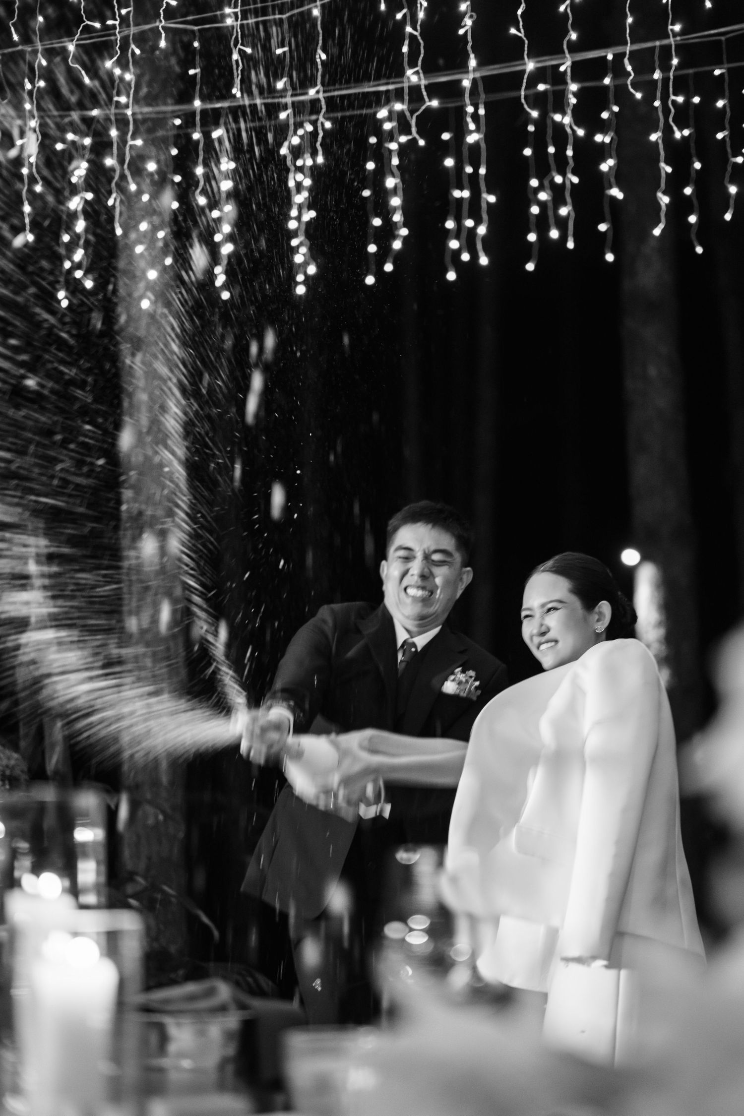 Black and white photo of laughing newlyweds popping champagne under sparkling fairy lights.