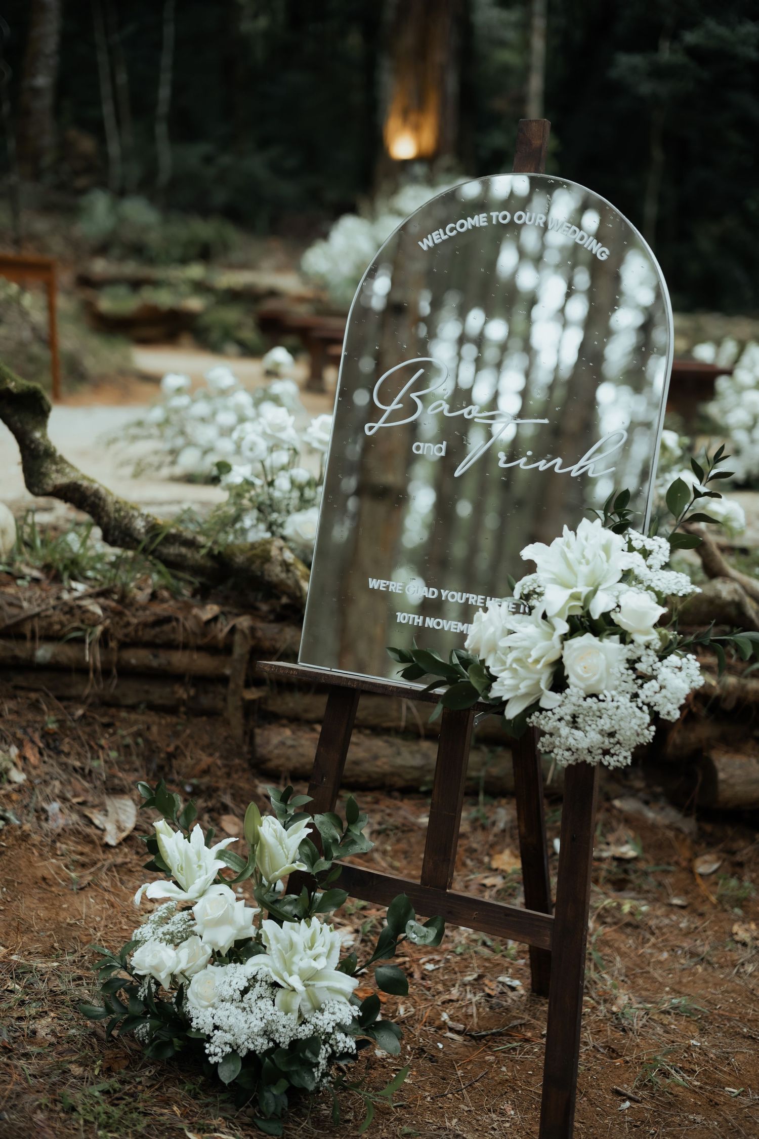 Elegant outdoor wedding welcome sign with white floral arrangements on wooden easel in forest.