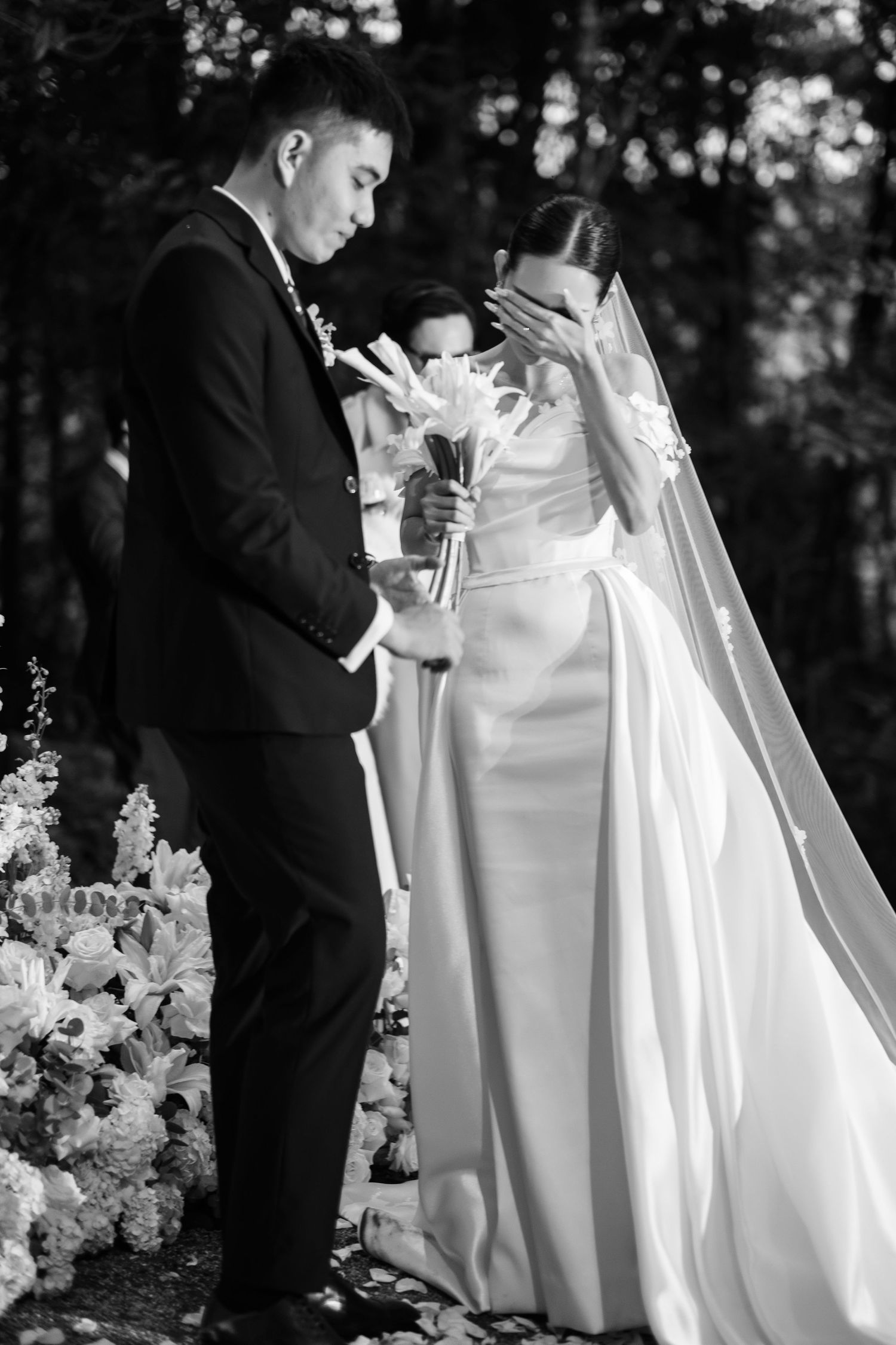 Emotional black and white photo of bride covering face while groom reads vows at forest wedding.