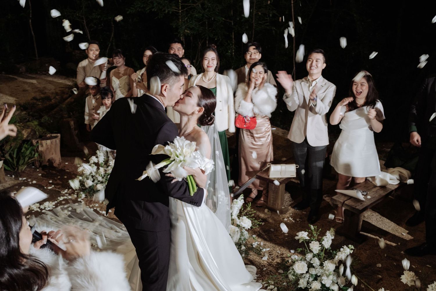 Joyful newlyweds celebrate first kiss as guests toss white petals at outdoor night wedding.