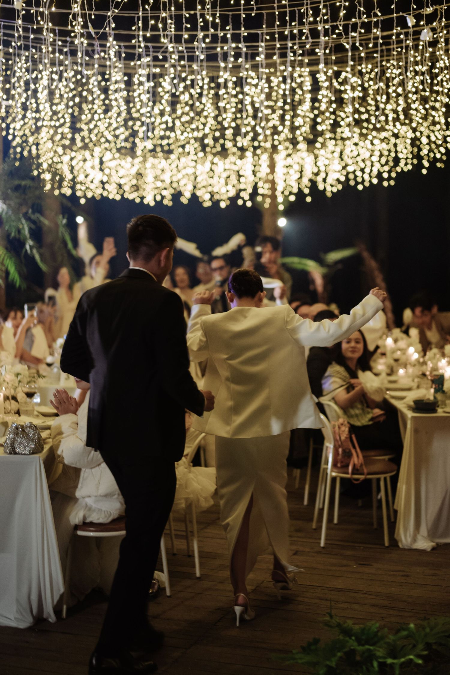 Bride and groom make grand entrance into outdoor fairy light reception as guests cheer.