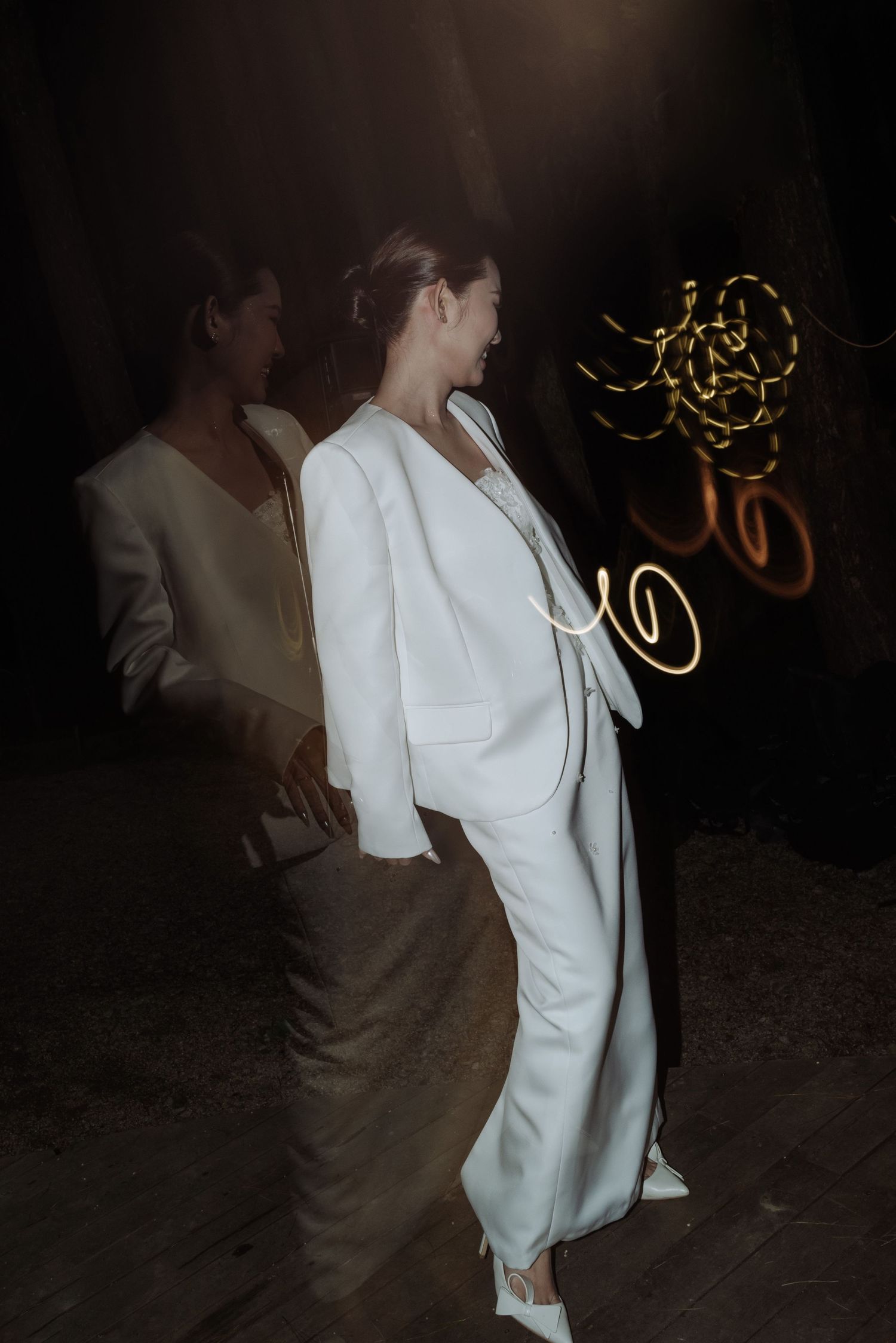 Bride in elegant white pantsuit smiling at an outdoor evening wedding reception with glowing light art installation.