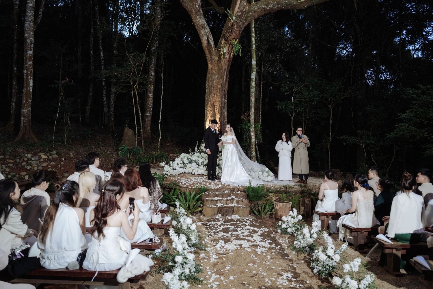 Intimate outdoor forest wedding ceremony at night with white floral aisle and seated guests.