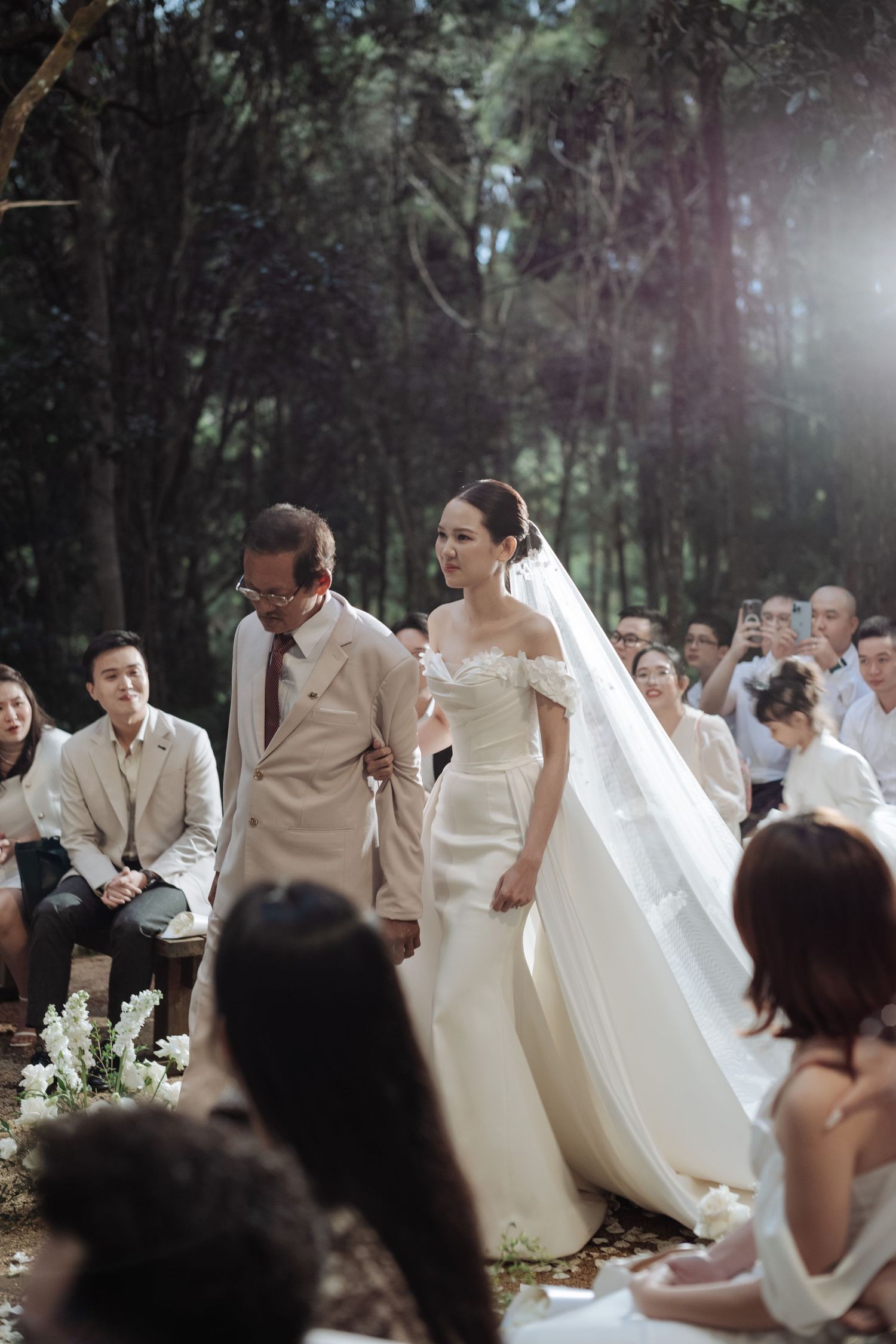 Bride in off-shoulder mermaid wedding gown walking down the aisle with escort in a sunlit forest ceremony.