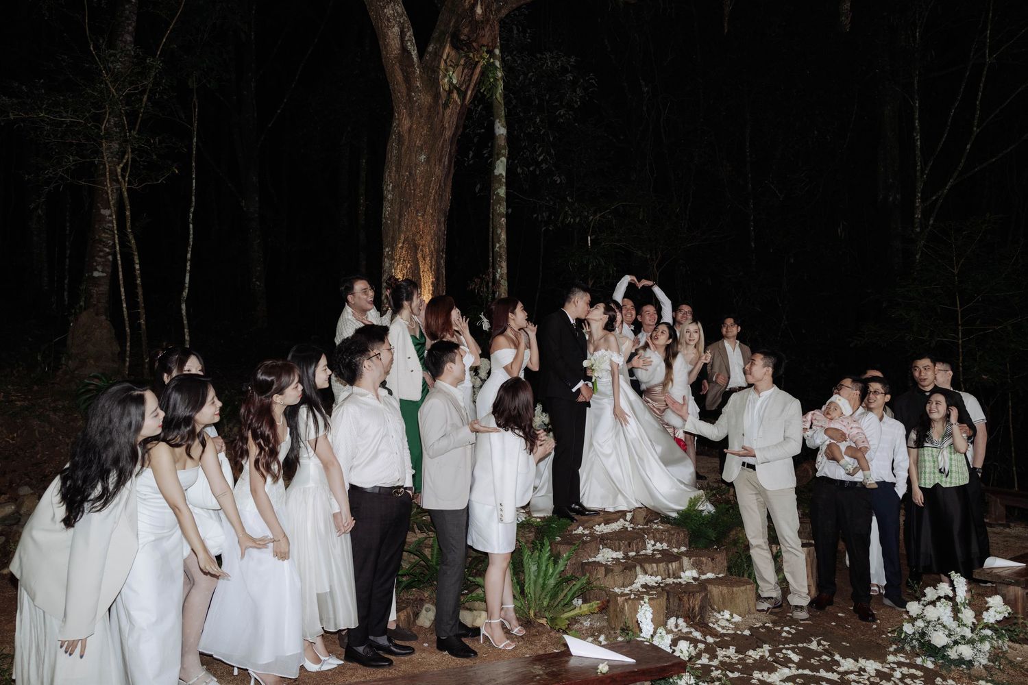 Wedding party group photo with couple at center raising hands in forest at night.