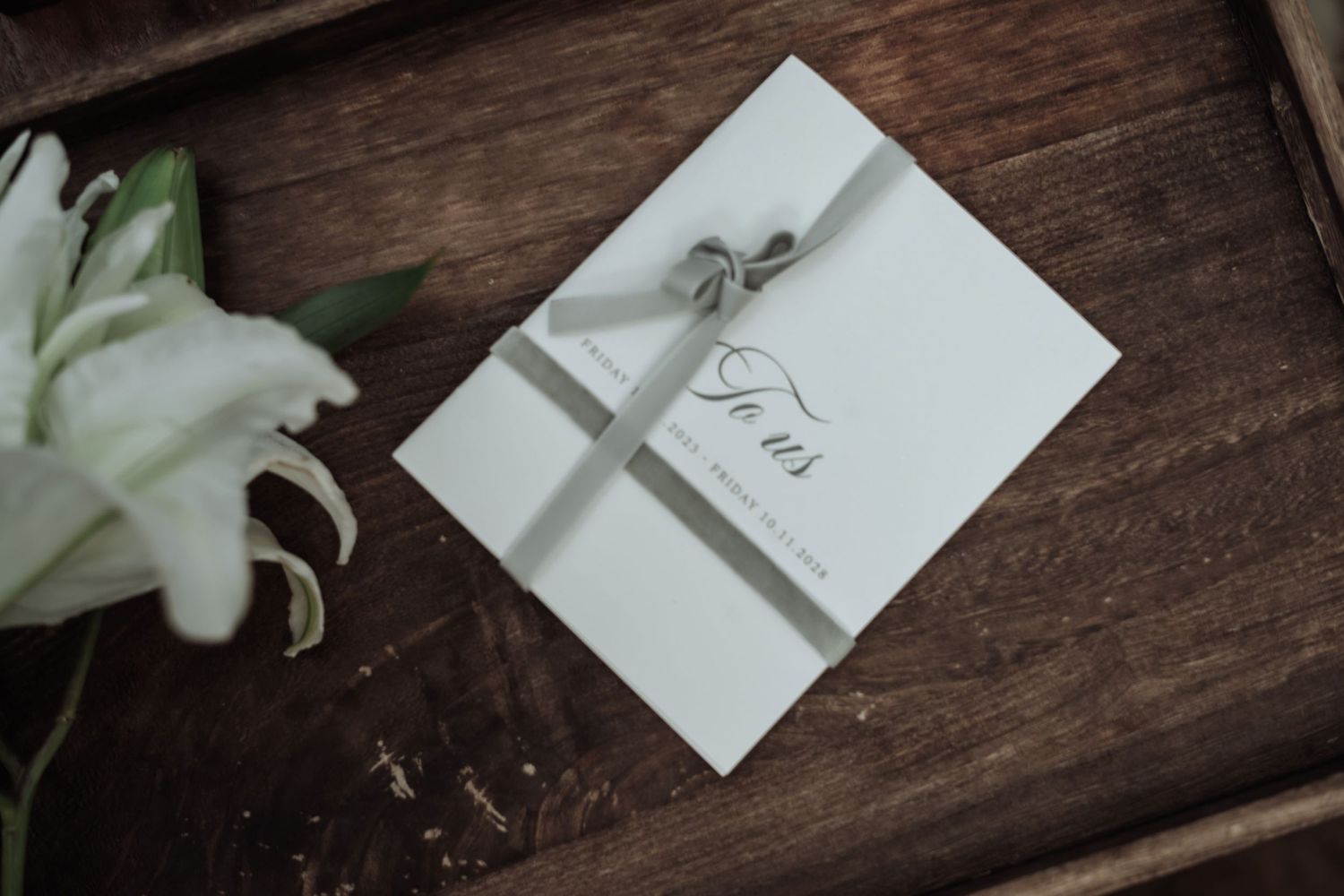 White wedding invitation card tied with ribbon beside white lily flower on wooden surface.
