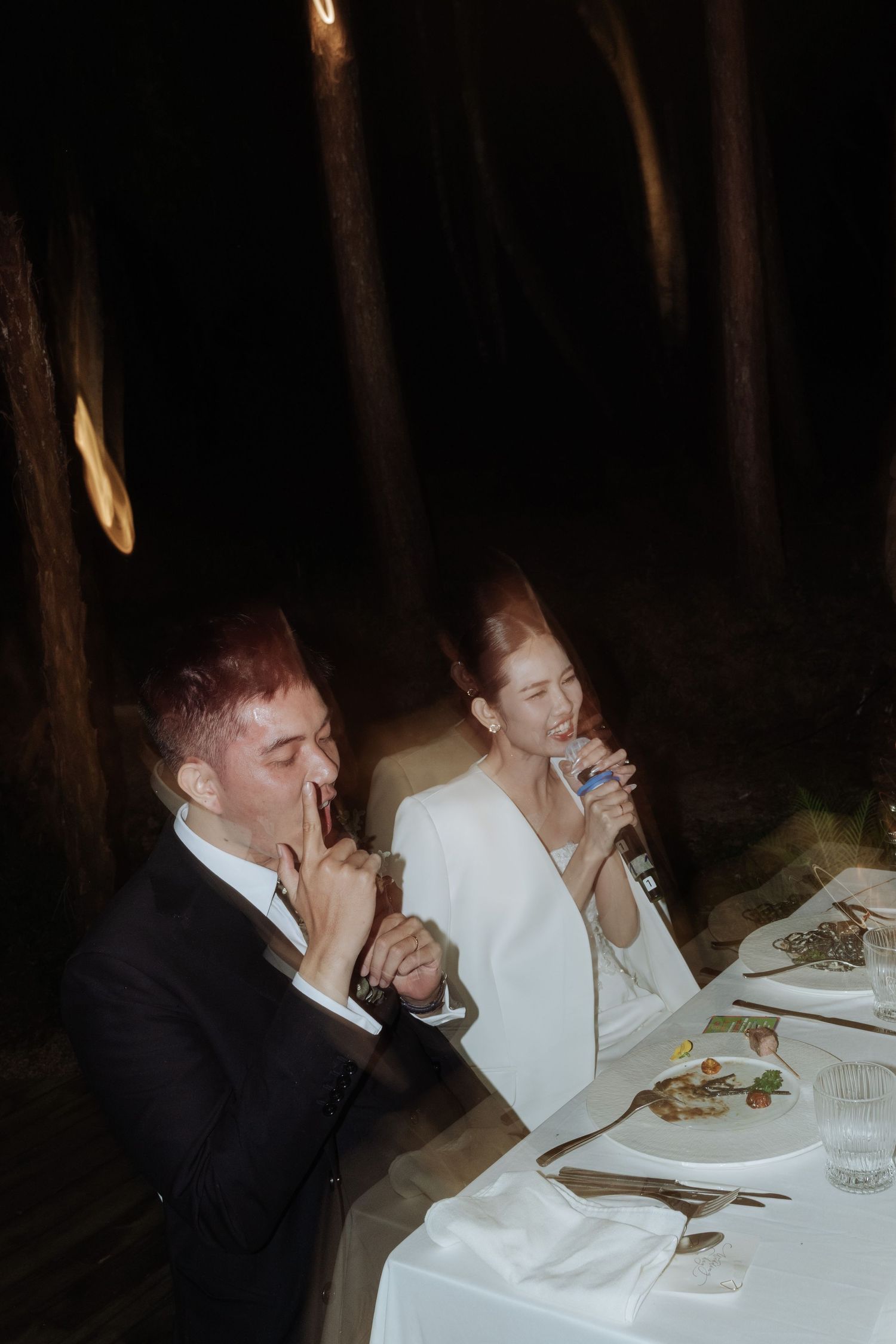 Bride and groom sharing drinks and laughing together at candlelit outdoor wedding reception table.