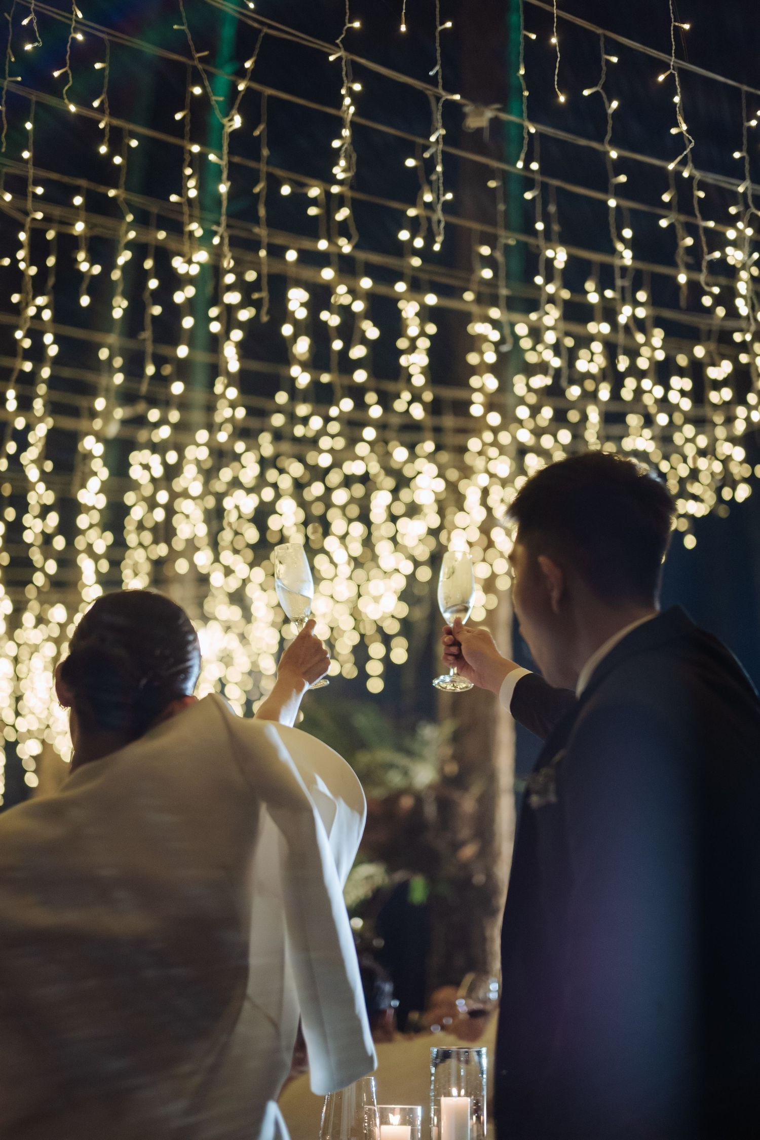 Bride and groom raising champagne glasses in toast under glowing fairy light canopy at night.