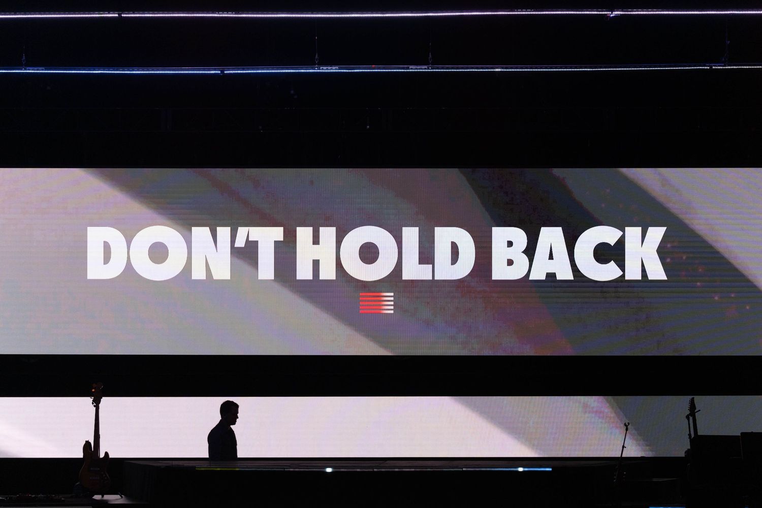 A large screen displaying the text "DON'T HOLD BACK". A person silhouette and a guitar can be seen in front of the scree