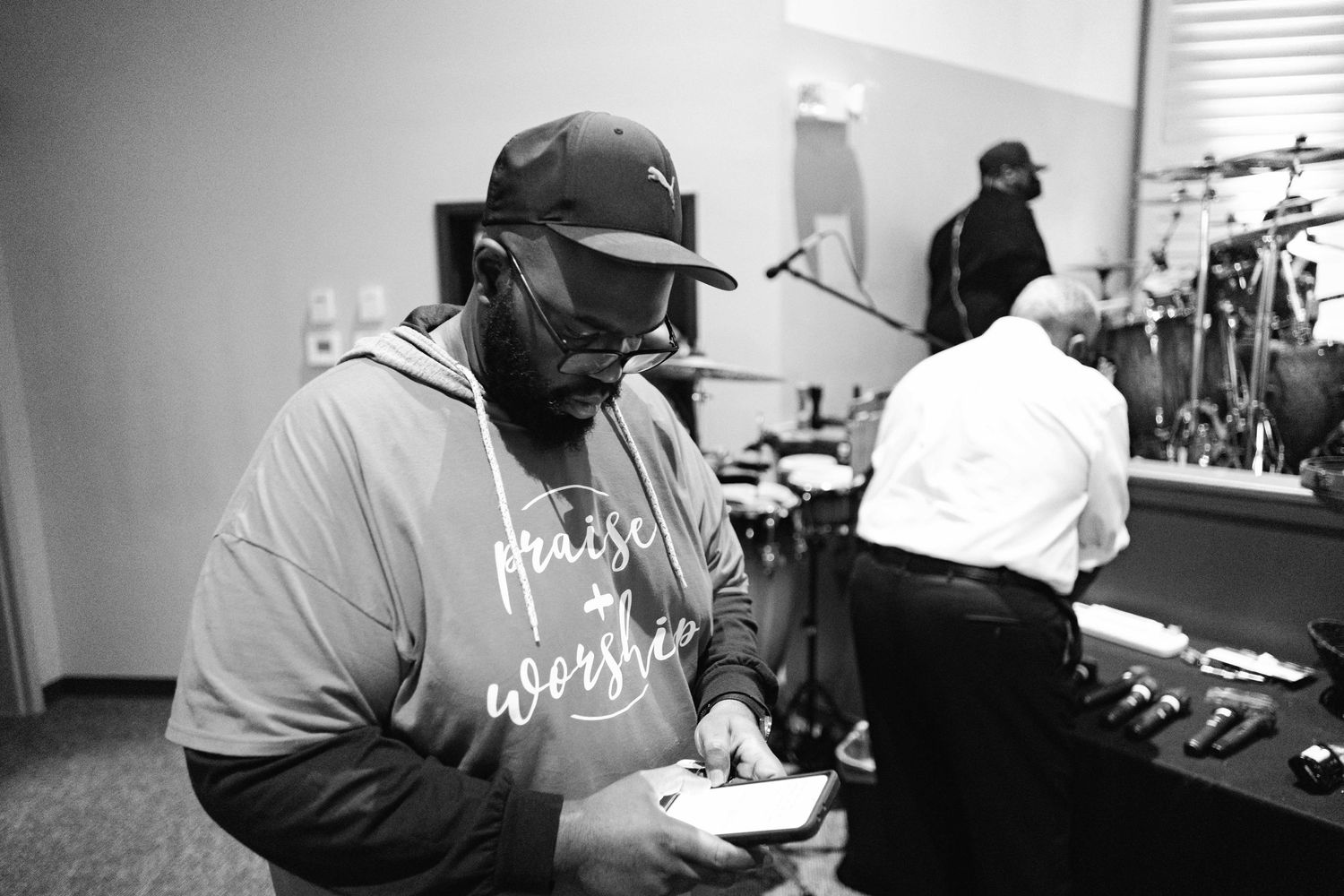 Black and white photo of a man wearing glasses and a cap, looking at his phone. His shirt reads "praise + worship." In t
