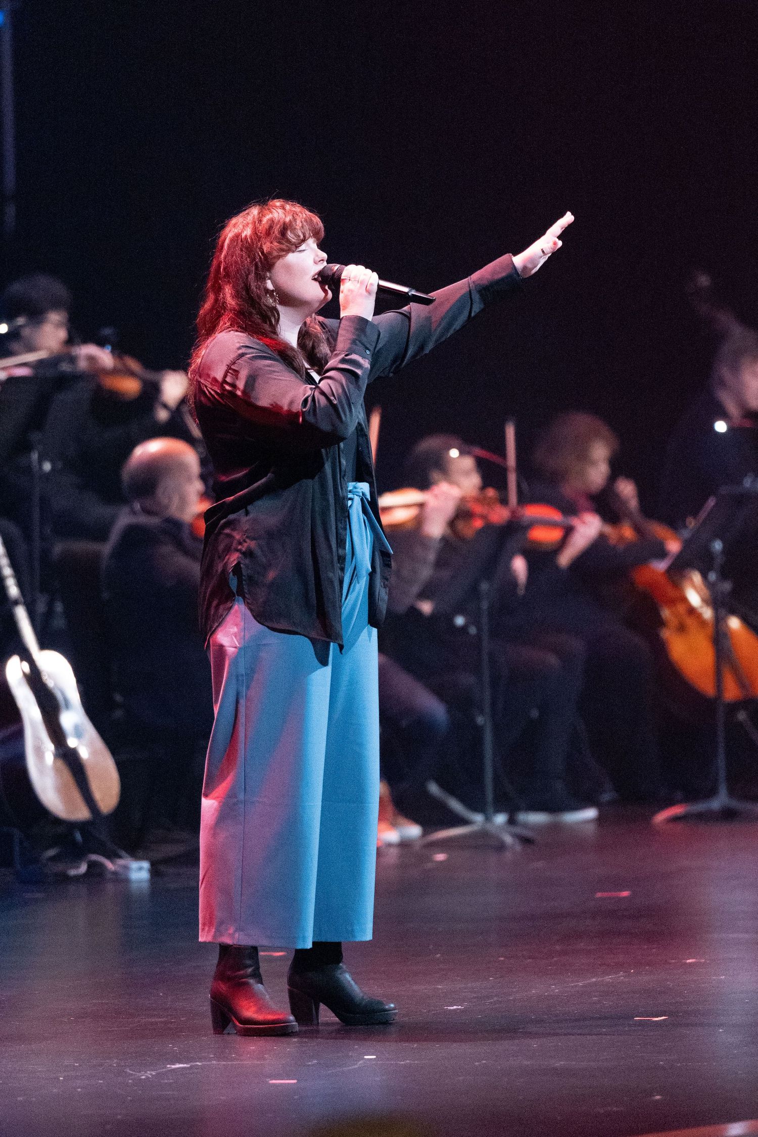 A woman is singing on stage with her right arm raised, wearing a black top and blue pants. She is performing in front of