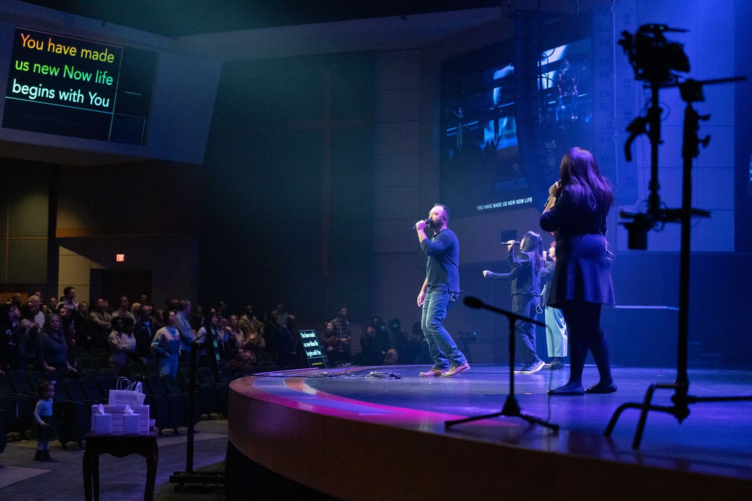 A group of people on stage performing music in front of an audience in a dimly lit auditorium. One person sings into a m