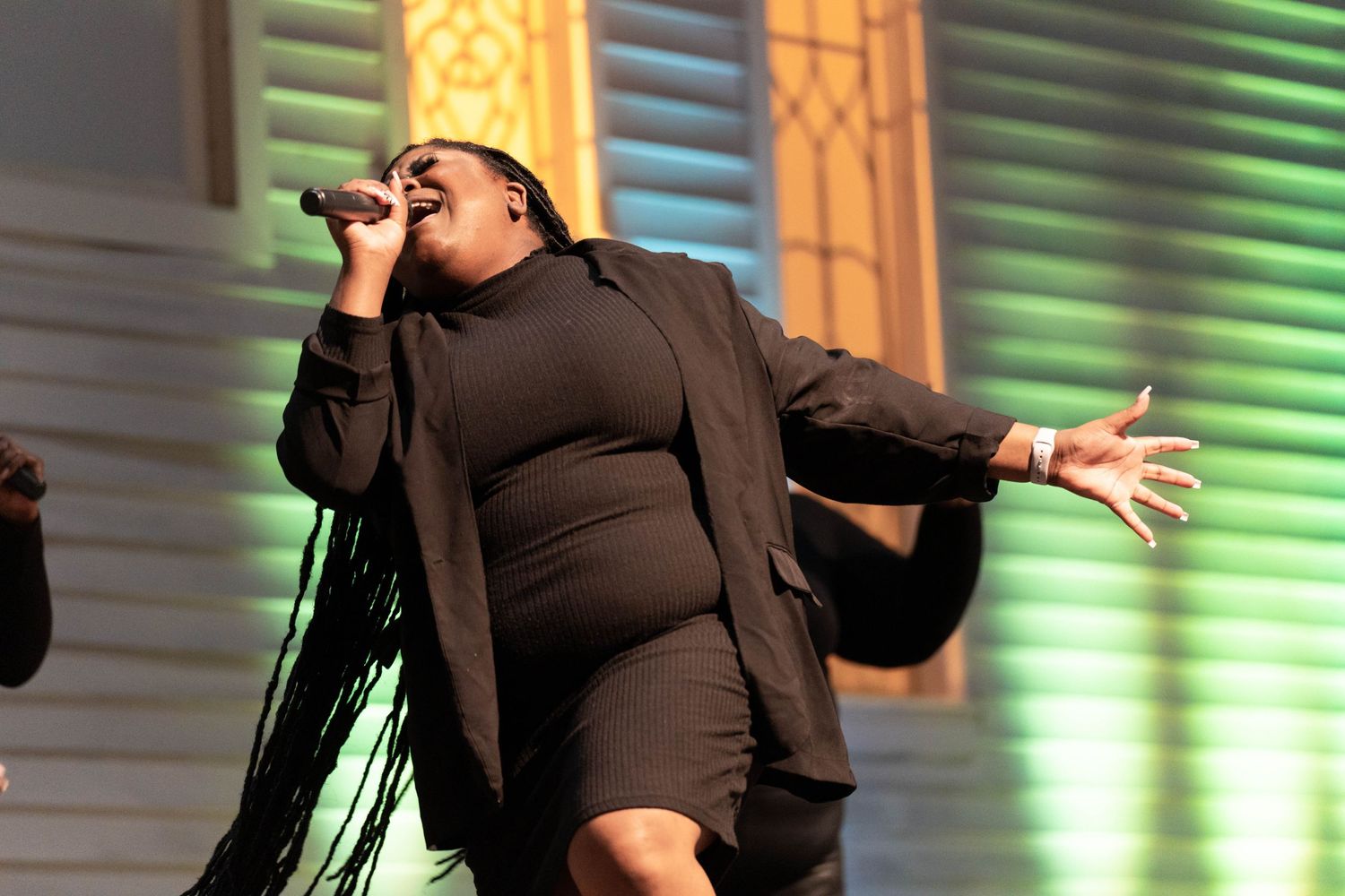 A person wearing a black outfit passionately sings into a microphone on stage. They have long hair and outstretched arms
