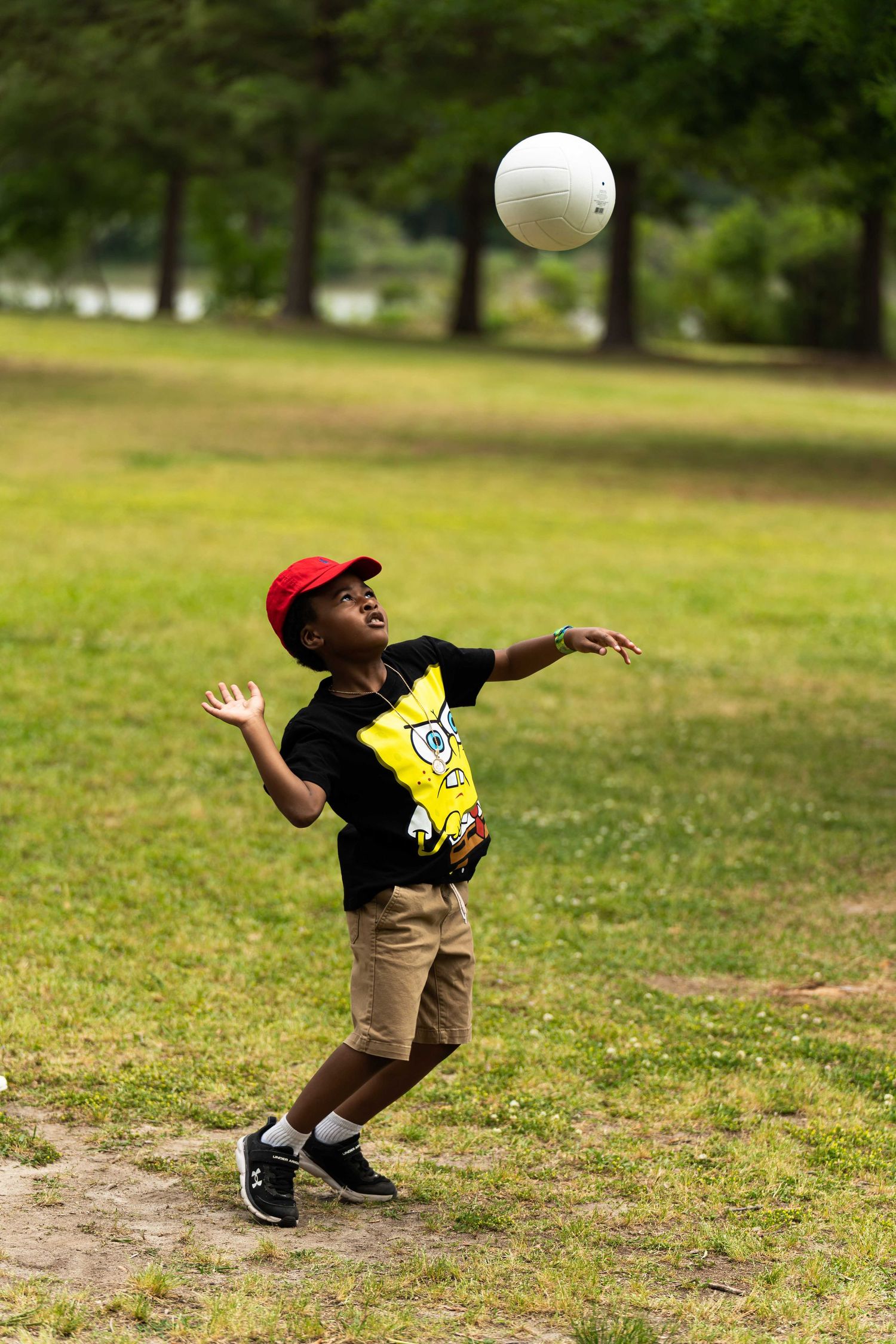 A young child wearing a red cap, SpongeBob t-shirt, beige shorts, and black sneakers is playing volleyball in a grassy p