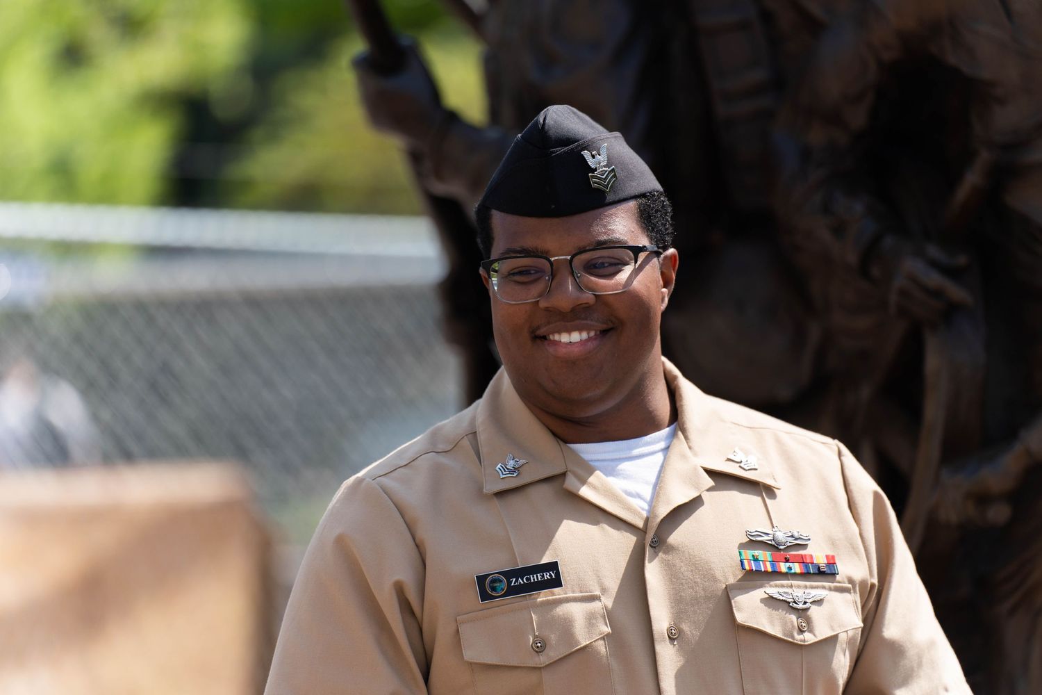 A person in a beige military uniform with a black beret and glasses is smiling. A name tag and various ribbons are displ