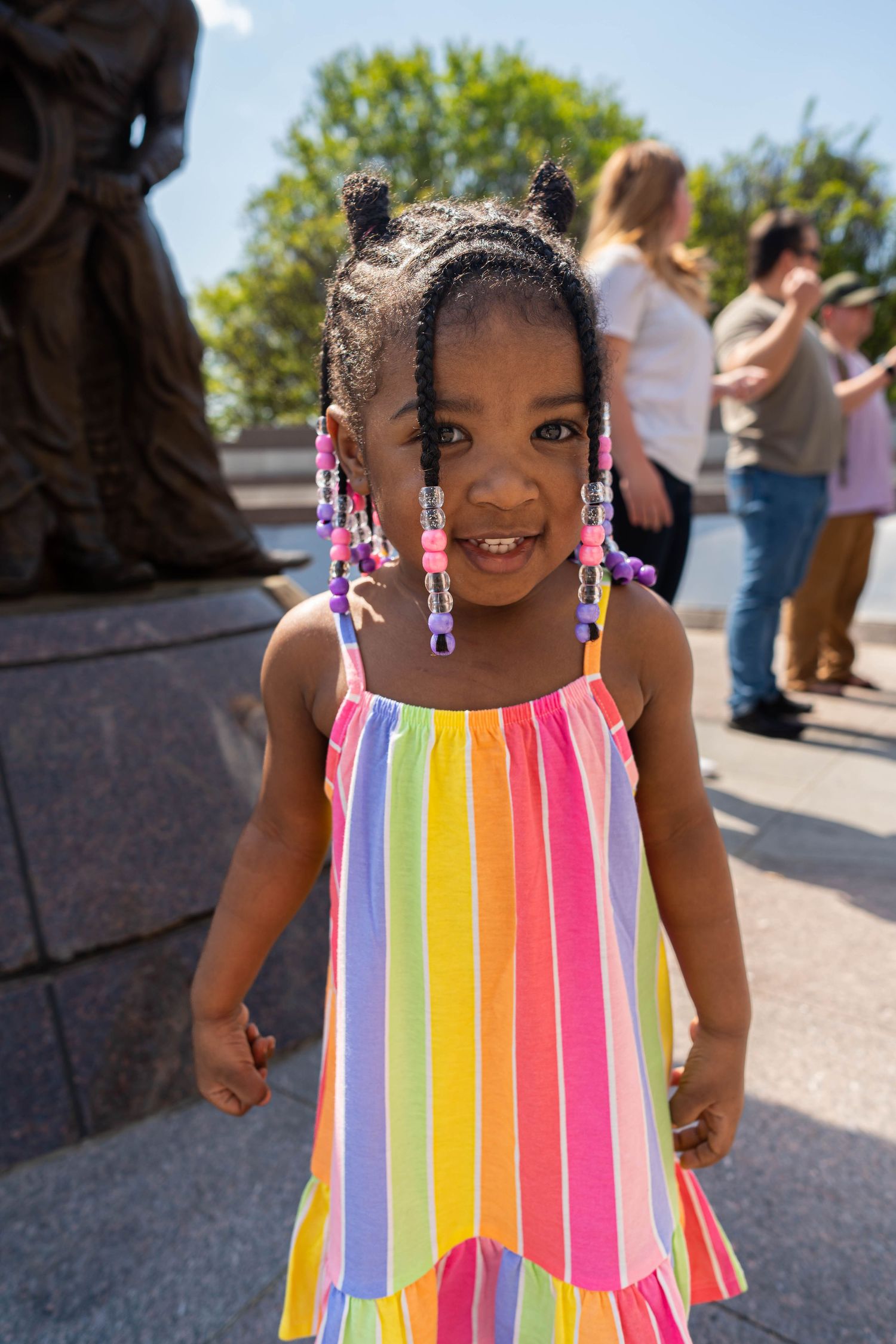 A young girl with beaded braids and a colorful striped dress smiles at the camera. She is outdoors on a sunny day with t