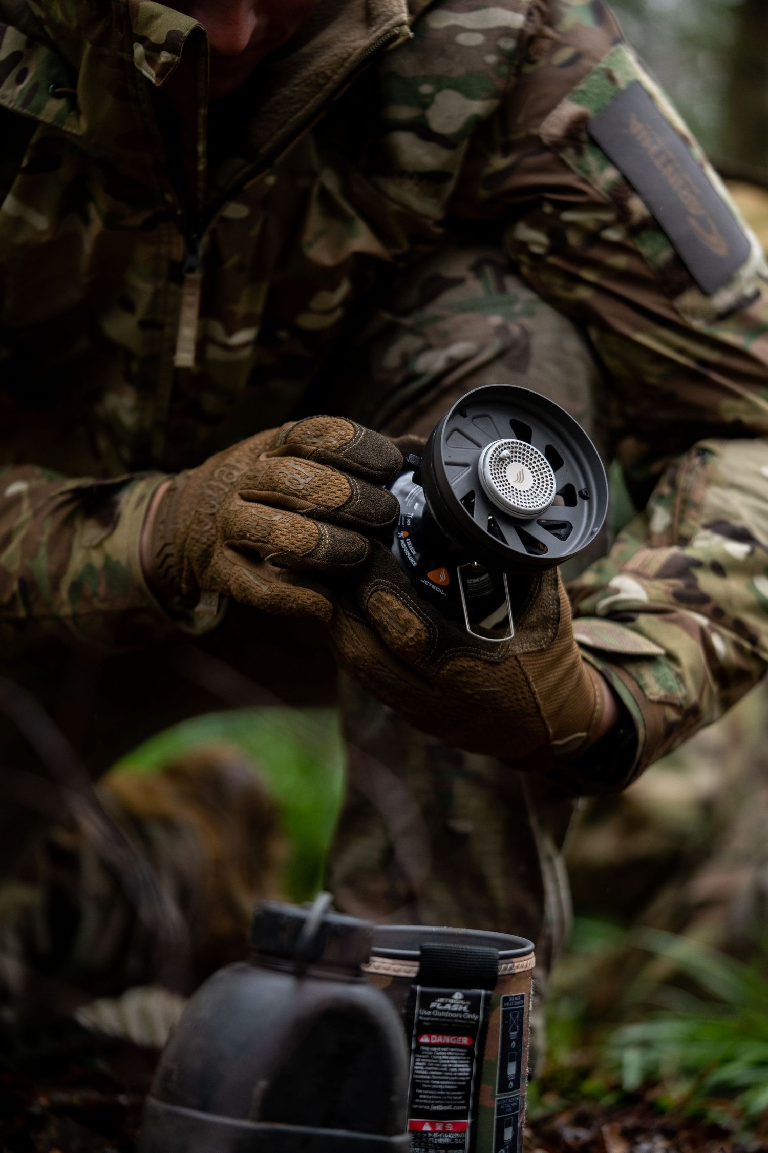 Camouflaged hands adjust camping stove burner in woodland setting.