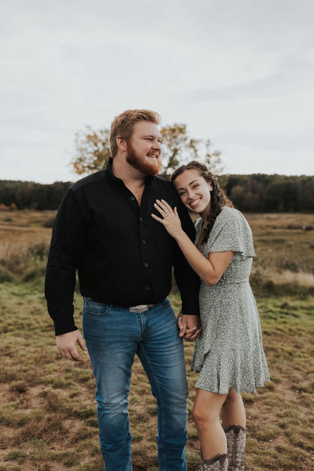 A romantic couple embraces in a rustic field, with her wearing a floral dress and boots while he wears jeans and dark shirt.