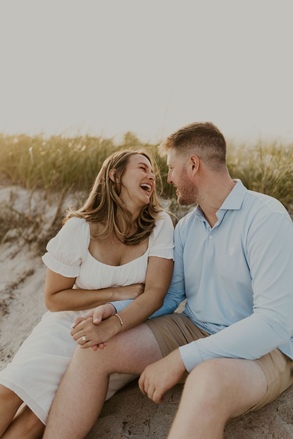 Couple sitting on sandy dunes sharing a genuine laugh during golden hour.