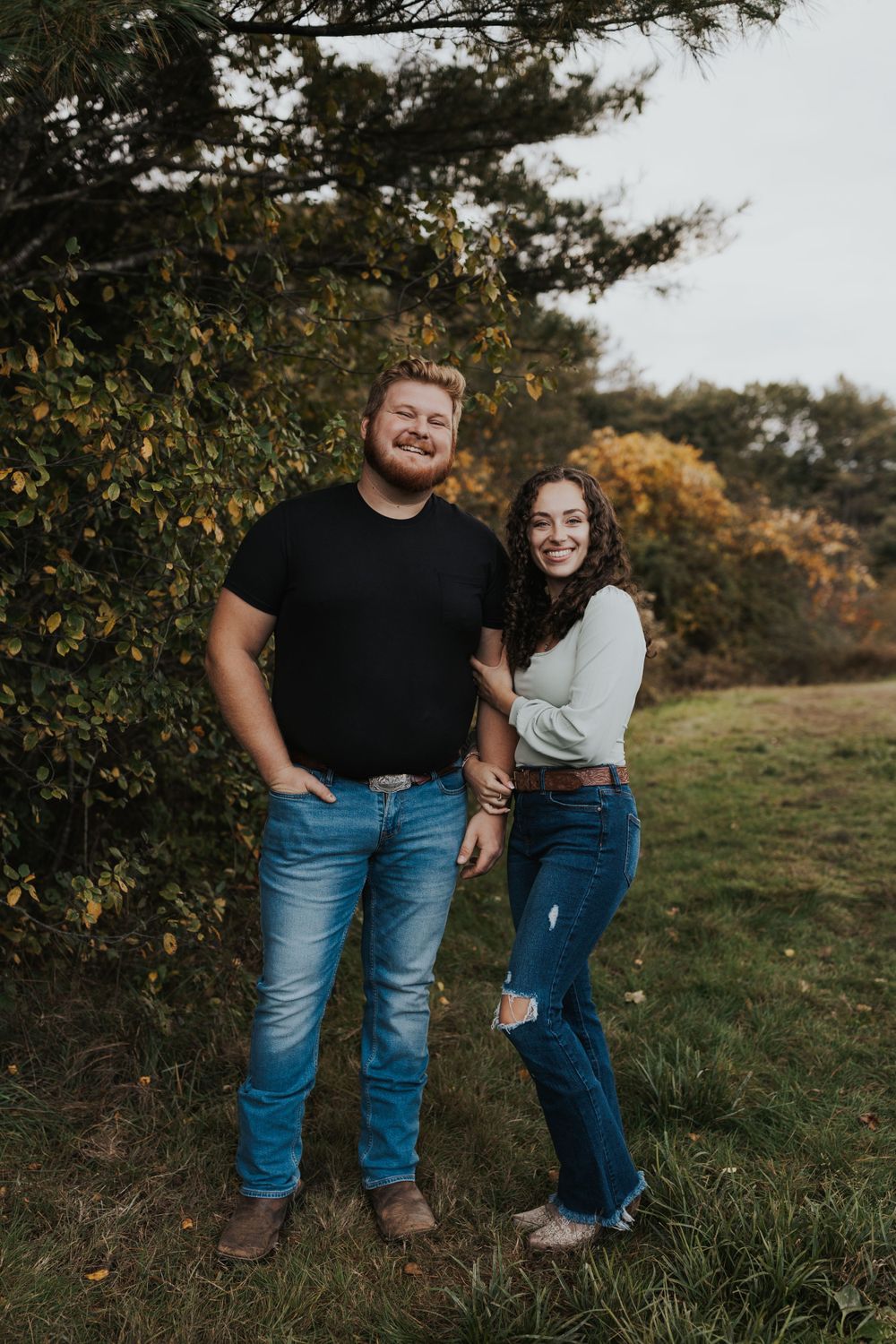 A joyful couple poses together against autumn trees in casual attire.