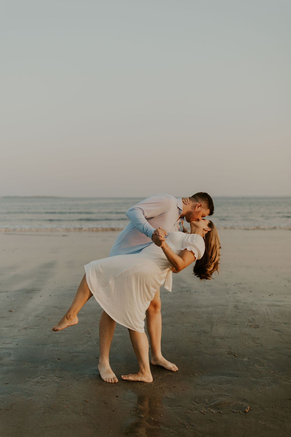Couple in light colored clothing sharing an intimate dip pose on a beach at dusk.