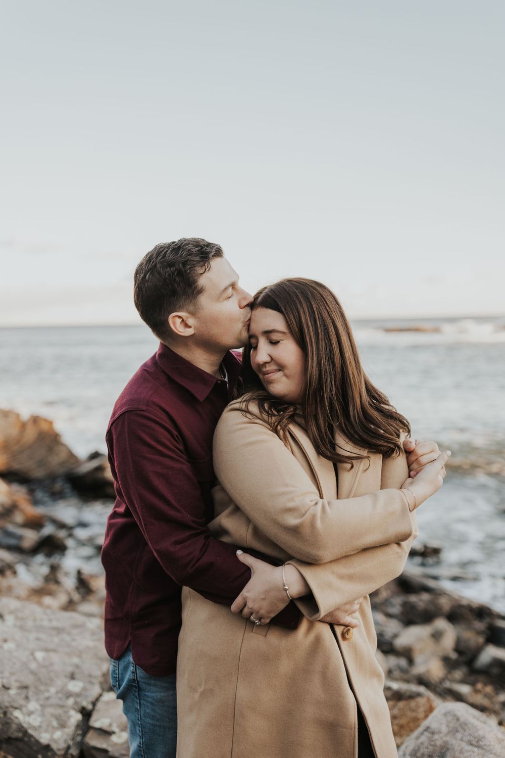Intimate moment between couple on rocky beach during coastal engagement session.