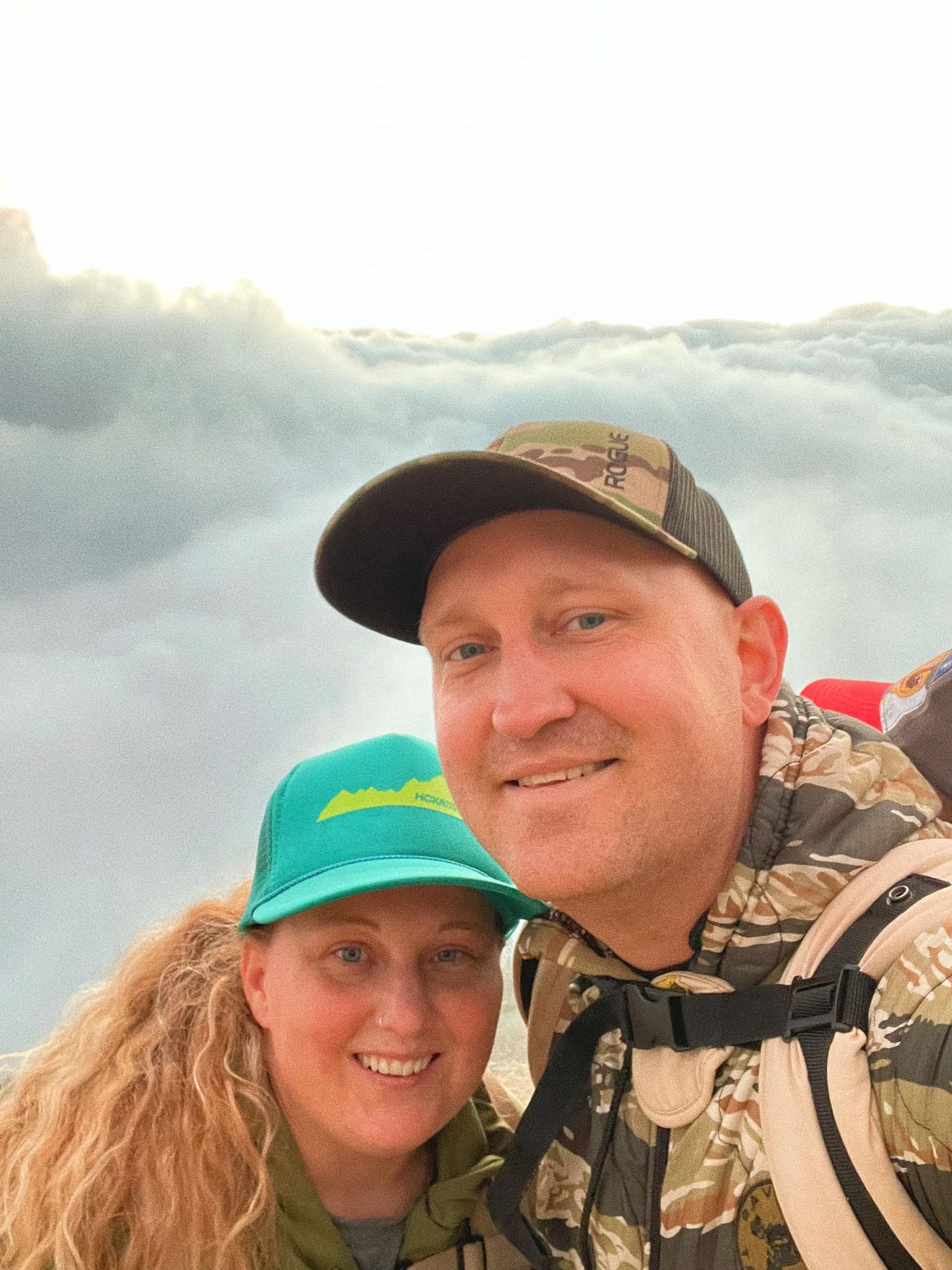 Two people wearing baseball caps smile together while hiking outdoors with cloudy skies in the background.